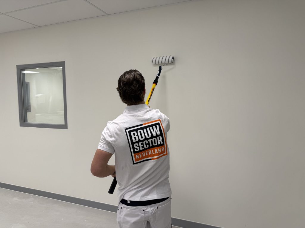 Schilder met Bouwsector-logo op zijn rug rolt een lichte wand naast een binnenraam met grijze lijst