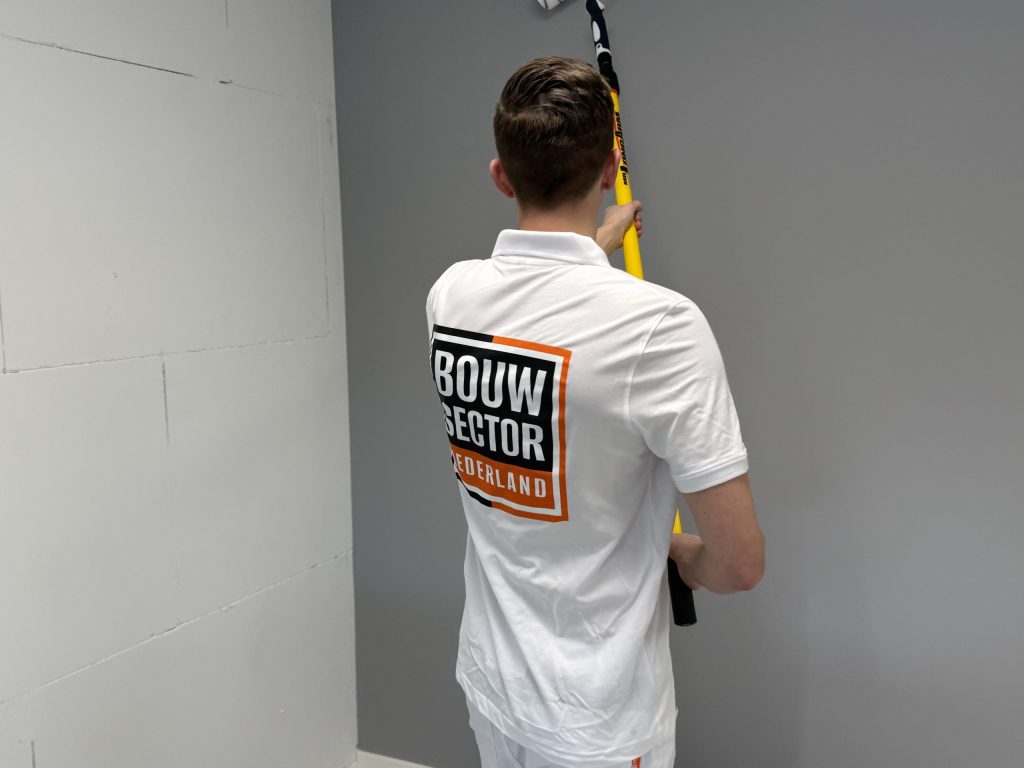Schilder in wit poloshirt van Bouwsector Nederland rolt een grijze muur naast een kale wand van kalkzandsteen