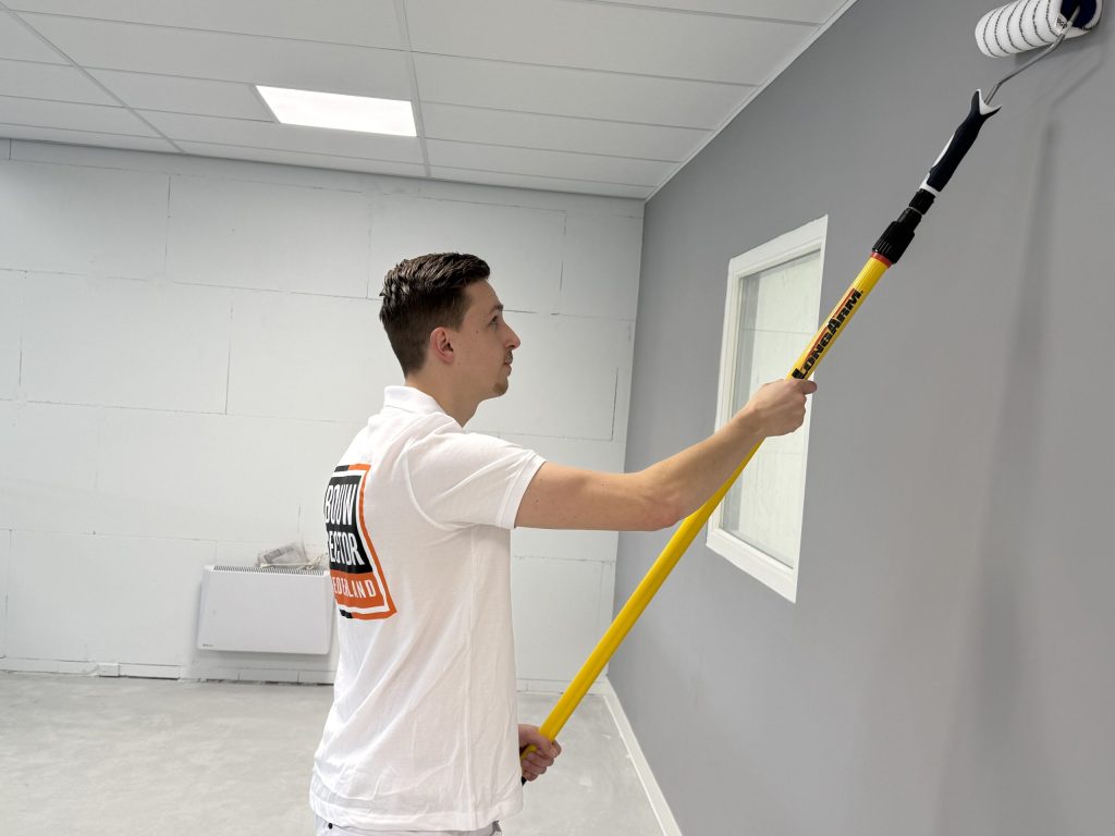 Schilder rolt een grijze wand naast een wit binnenraam in een lege ruimte met systeemplafond
