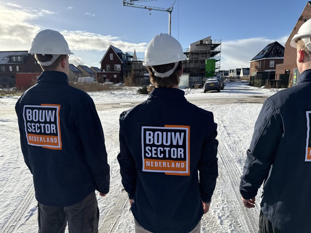 Drie mannen in werkjas met het logo van Bouwsector Nederland voor een rij huizen onder een blauwe lucht