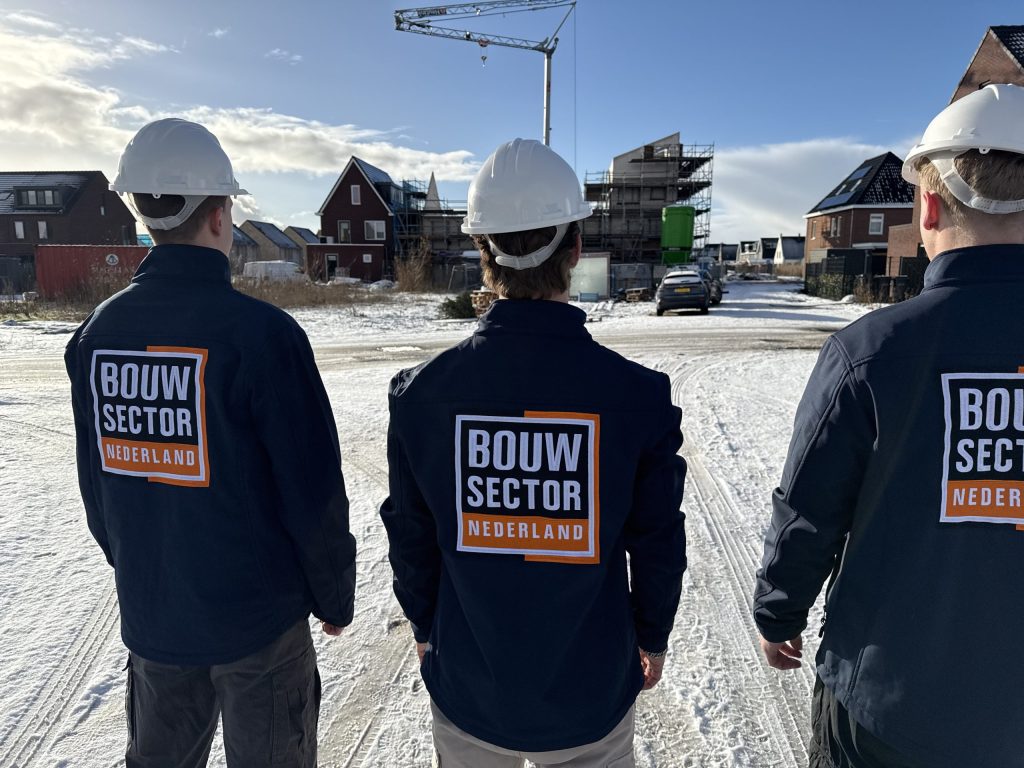 Drie medewerkers van Bouwsector Nederland in werkjas met logo en witte helm op een besneeuwde straat met nieuwe woningen
