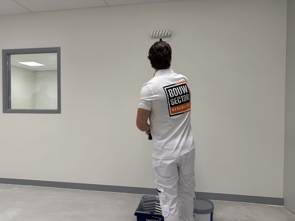 Schilder in een wit poloshirt verft een lichte muur naast een binnenraam met een grijs kozijn